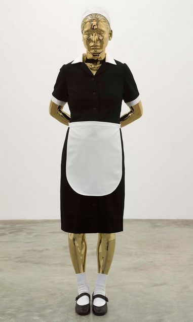 Eine goldfarbene Puppe steht vor einem neutralen Hintergrund. Sie hat die Hände hinter dem Rücken verschränkt und trägt eine Hausmädchenuniform mit einem schwarzen Kleid, einer weißen Schürze, weißen Socken und schwarzen Schuhen. Ihr Blick ist leicht nach unten gerichtet.