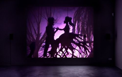 Against a purple background, a black silhouette stands out. A woman and a man are dancing in front of sparse, gnarled trees.