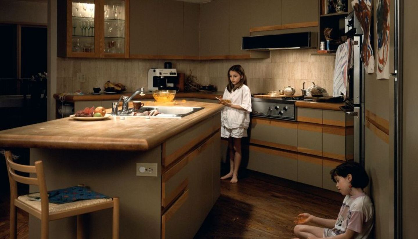 Zu sehen ist eine geräumige Küche mit einer zentral platzierten Kücheninsel. Am rechten Bildrand sitzt ein Mädchen im Schneidersitz auf dem Boden und lehnt an einem Kühlschrank. Ihre Handbewegung vermittelt den Eindruck, als würde sie gerade etwas erklären oder erzählen. Hinter der Kücheninsel steht ein weiteres Mädchen, das sich an den Herd anlehnt. Ihr Blick ist in den Raum gerichtet, während ihre Haltung nachdenklich oder gedankenverloren wirkt.