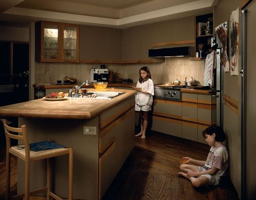 Zu sehen ist eine geräumige Küche mit einer zentral platzierten Kücheninsel. Am rechten Bildrand sitzt ein Mädchen im Schneidersitz auf dem Boden und lehnt an einem Kühlschrank. Ihre Handbewegung vermittelt den Eindruck, als würde sie gerade etwas erklären oder erzählen. Hinter der Kücheninsel steht ein weiteres Mädchen, das sich an den Herd anlehnt. Ihr Blick ist in den Raum gerichtet, während ihre Haltung nachdenklich oder gedankenverloren wirkt.