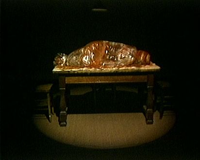 In einem dunklen Raum beleuchtet ein Scheinwerfer einen Tisch. Darauf liegt etwas in durchsichtiger Folie verpackt, veilleicht eine Person. Rote Farbe scheint durch die Folie durch und erinnert an Blut.