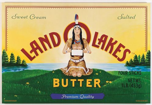 Ein rechteckiges, großformatiges Bild ist im Stil einer Butterverpackung gestaltet. In roter Schrift auf gelbem Hintergrund steht der Markenname „Land O Lakes“, darunter das Wort „Butter“ vor einer grünen Wiese. In der Bildmitte sitzt eine Frau mit langen dunklen Zöpfen in Kleidung, die an traditionelle Bekleidung indigener nordamerikanischer Völker erinnert. Das Bild enthält eine Klappmechanik im Brustbereich der Figur, durch die beim Öffnen der Oberkörper freigelegt wird.