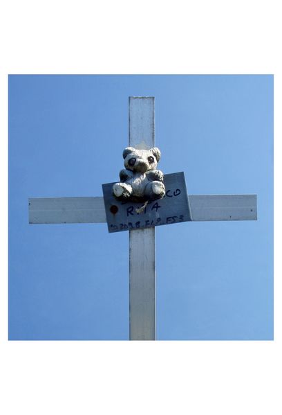Photographs of white grave crosses against blue cloudless sky. All have simple memorial plates made of sheet metal, on which the names and life dates of the deceased are handwritten. Children's toys or stuffed animals are attached to many of the crosses. Both the crosses and the toys have been soiled by the weather and have come down. In some photos, wild grasses and yellow flowers protrude into the picture from below. 