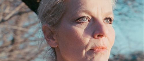 Close up on the face of a woman with white, tied up hair. In the background is blue sky and tree branches