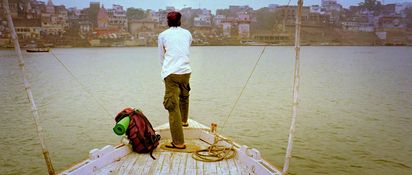 Rückenansicht von einer Person, die auf einem Schiffbug steht. Neben ihm liegt ein Rucksack, vor ihm ist ein großes Gewässer und im Hintergrund Festland mit einer Stadt.
