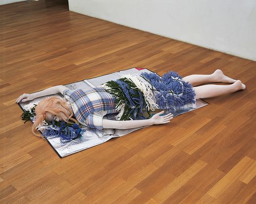 Eine Puppe liegt mit dem Kopf dem Boden zugewandt auf einer silbernen Isolationsmatte. Sie trägt einen Rock mit applizierten Wollfransen und ein kariertes Oberteil. Ihre Haare sind erdbeerblond.