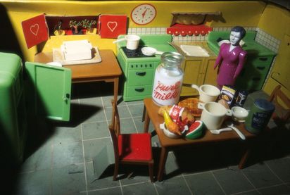 er gelb gestrichene Küchenraum eines Puppenhauses wurde schräg von oben fotografiert. In der Ecke der Küche steht eine Puppenfigur in einem lilafarbenen, glänzenden Kleid. Auf dem Tisch sind verschiedene Lebensmittel ausgebreitet. Besonders auffällig ist ein Milchgefäß mit der roten Aufschrift „Pure Milk“.