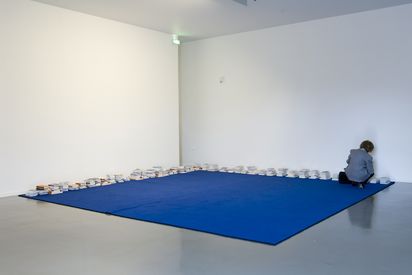 A large square blue carpet lies on the floor in the corner of the room. Stacks of books lie on the carpet along the walls. A woman is kneeling and reading one of the books 