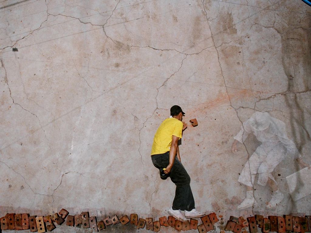 Vor dem Hintergrund einer großen Steinplatte stehen zwei Figuren auf einer Reihe aneinandergereihter Ziegel – zumindest sieht es so aus. Das Foto wurde jedoch von oben aufgenommen, und die Figuren liegen tatsächlich. Eine Person ist in Weiß gekleidet und scheint mit einem Ziegelstein auf die in Gelb gekleidete Figur zu werfen.