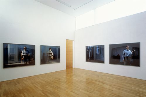 Four large-format photographs hang around a corner in an exhibition space. All the photos show a man in a dark apartment