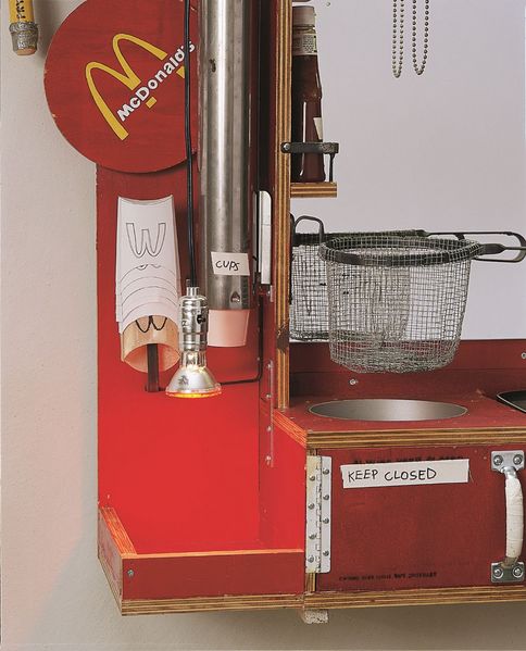 Bildausschnitt einer größeren im McDonalds-Stil gehaltenen Objektstruktur. Das Objekt setzt sich aus mehreren Alltagsgegenständen zusammen. In diesem Ausschnitt sieht man das Netz einer Fritteuse, einen händisch beschrifteten Becherhalter, Pommestüten mit handgemalten McDonalds-Zeichen sowie eine tiefgehängte kleine Lampe, die einer Wärmelampe ähnelt. 