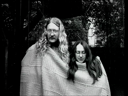 Ein Standbild aus einem filmischen Werk, bei dem zwei Personen zusammen in das selbe große Stück gestrickten Stoff gehüllt sind.