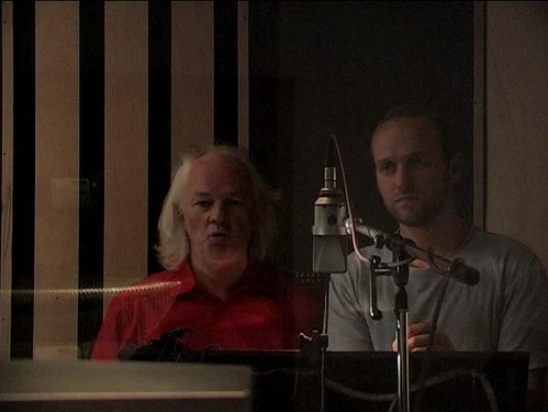 In a recording studio, two men stand next to each other in front of a microphone. One has chin-length white hair, wears a red shirt and looks like he's whistling into the microphone. 