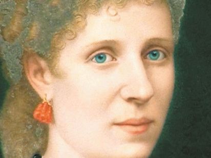 Half profile of a woman with light blue eyes and reddish blond curly hair. She wears a coral coloured earring.