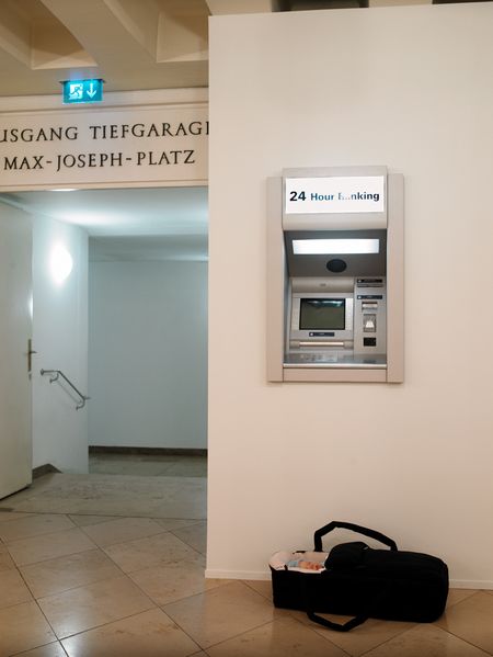 Vor einem Tiefgaragenzugang ist ein Geldautomat in eine Wand eingelassen. Darüber befindet sich eine weiße Leuchttafel mit der Aufschrift „24-Hour Banking“. Vor dem Automaten steht eine schwarze Tragetasche, in der ein kleines Kind liegt.