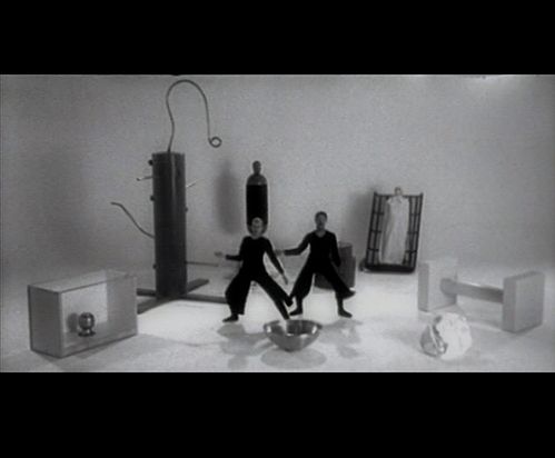 Black and white video screenshot: a kind of stage with objects that cannot be clearly identified. In the middle are two dark-clad figures who look as if they are dancing