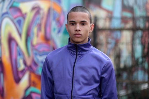 A young man with short-cropped hair and a purple jacket stands in front of a graffiti wall