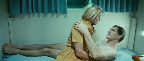 In a ship's cabin, a naked man lies on the bed and pulls a woman sitting on the edge of the bed towards him.