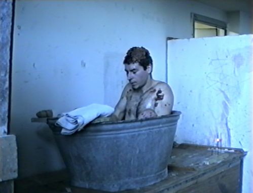 A man sits in a small metal tub. A brown mass sticks to his naked upper body and head