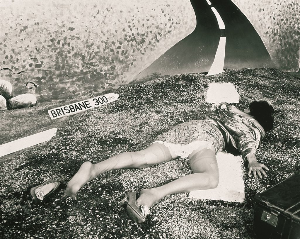 Black and white photograph of a woman in a dress with a snake pattern lying on the ground in the middle of a road as if she had been run over. Her dress has ridden up and she has lost a shoe; a suitcase lies next to her. A road sign reads “Brisbane 300 miles”.