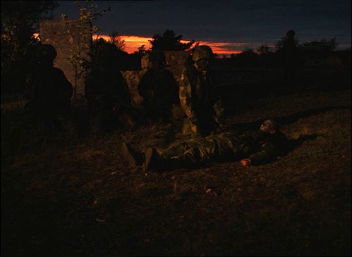 Very dark scene at dusk. A person in a military camouflage suit is lying on the ground outside. Other people in military uniforms are standing around them.