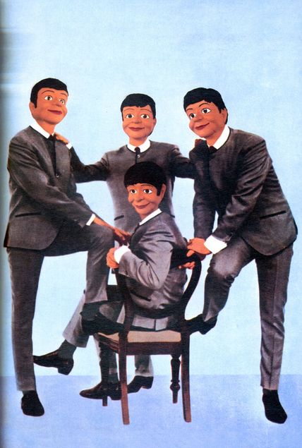 Four ventriloquist dummies with black bob hairstyles are dressed in grey suits. Three of the figures stand in front of a blue background. The fourth sits on a chair, which the others are lightly leaning against.