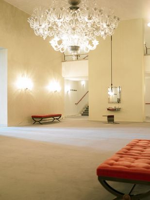 In the white-painted foyer of an opera house, red-upholstered benches line the walls. On a wall without a bench hangs a mirror framed with light bulbs. In front of it lies an overturned chair. From the ceiling hangs a looped rope.