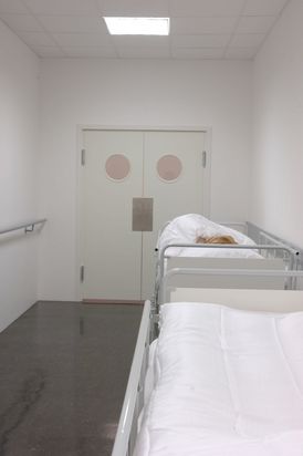 A figure with light blond hair lies in a hospital bed, their head turned toward the wall. The image shows the upper body wrapped in a white blanket. The face of the waxen-looking figure is only partially visible.