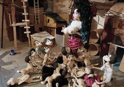 Eine Knetfigur-Frau steht vor einer Schar Knetfigur-Kinder, die sich vor ihren Füßen am Boden tummeln. Die Frau hat schwaze lockige Haare, einen blassen Teint und stark geschminkte Augen und Lippen. Ihre Bluse ist vorne geöffnet und sie hält die Brüste den Kindern entgegen. Aus der einen Brust tropft eine weiße Flüssigkeit. DeKinder, alle mit kurzen dunklen Haaren, schauen zu der Frau hoch, als würden sie sie anflehen. 