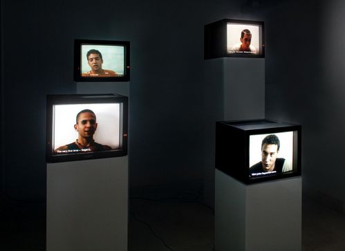 Four monitors on four plinths of different heights. All of them show men's heads talking into the camera.