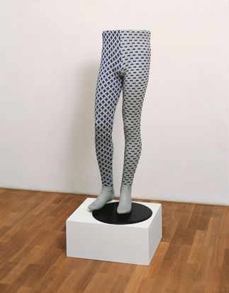Skulptur eines Körperunterteils ohne Oberteil, stehend auf einem Sockel; die Beine tragen blau-grau gemusterte Strumpfhosen, das eine Bein trägt ein Plus-Muster, das andere ein Minus-Muster.