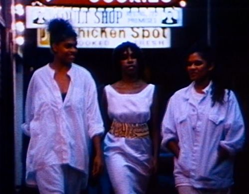 Three black women in white clothes walk side by side on the street, behind them are glowing advertising signs.