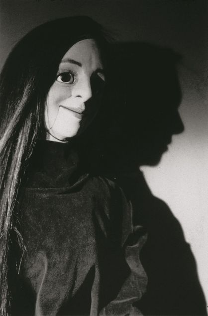 A ventriloquist dummy with a  movable lower jaw is depicted in a black-and-white photograph. It wears a dark-haired wig and a black robe.