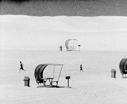 Schwarzwißaufnahme shows a moonscape-like area with individual people and covered picnic tables and benches, each with a garbage can and a barbecue.