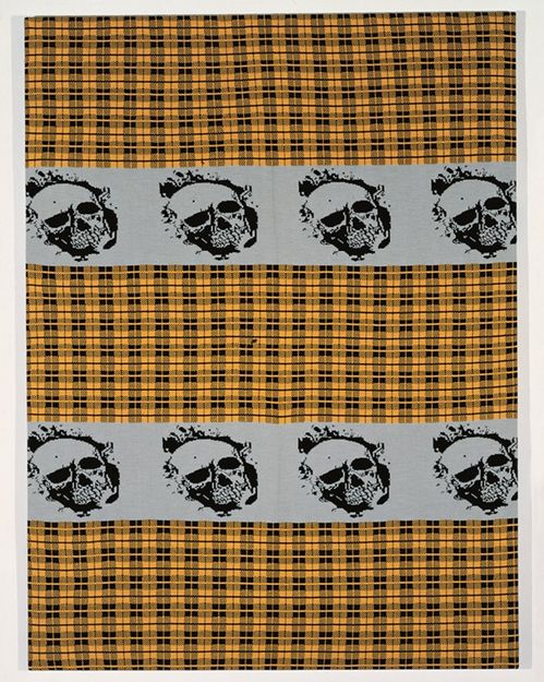 Ein quadratisches Strickstück mit schwarzem Karomuster auf ockerfarbenen Grund, zentral oben und unten zwei graufarbene Streifen mit einer sich wiederholenden schwarzfarbenen Totenkopfabbildung.