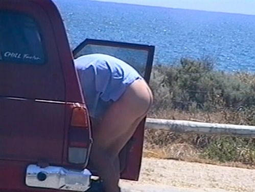 A person stands in front of a beach with his head bent into a car and his naked lower body outside. 