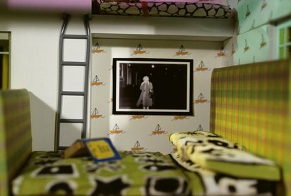 In einer Frontalaufnahme ist ein Zimmer eines Puppenhauses zu sehen, dessen Wände mit einer Tapete mit Segelschiffen versehen sind. Im Vordergrund, sich über den gesamten Bildraum erstreckend, steht ein grün kariertes Bett mit grün gemusterter Bettwäsche. Neben einer Leiter, die zu einem zweiten Bett hinaufführt, ist ein schwarz-weißes Bild angebracht, das eine blonde Frau zeigt, die ihren Mantel hochschlägt, um durch ein Schneegestöber zu gehen.
