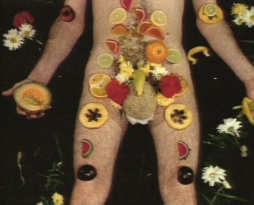 Video screenshot: a naked male body is covered with flowers and fruit