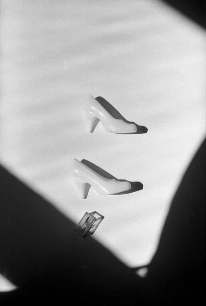 Two Barbie toy shoes are photographed on a white surface. A dark shadow forms a natural frame around the light-coloured shoes. The photograph is rendered in black and white.