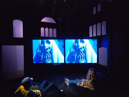 Two projections are played in front of a house backdrop, both showing the image of a ghost. Viewers sit in front of them on cushions.