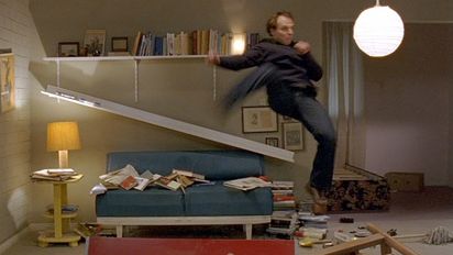 In a living room, a man hangs in the air as if he had jumped up. Behind him, a bookshelf has come loose from the wall and the books are lying on the couch and the floor.