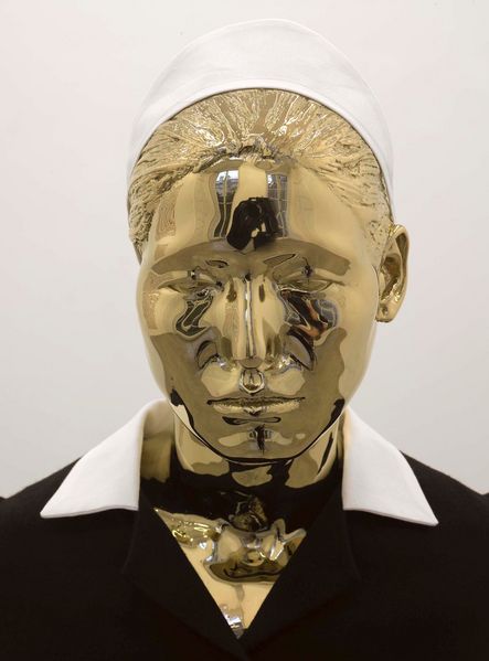 In dem goldfarbenen Gesicht einer Modellpuppe spiegelt sich leicht der umliegende Raum. Das Gesicht gehört zu einem Hausmädchen, wie man an dem Ansatz einer dunklen Uniform, einem weißen Kragen und einer gestärkten, weißen Haube erkennen kann.