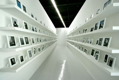 The image shows a section of a corridor with white shelves mounted on the walls. On the shelves are photographs in white frames. The photographs mainly depict young people, mostly male couples.