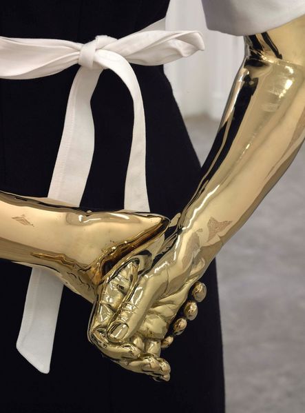 Man sieht die verschränkten goldfarbenen Hände einer Modellpuppe, die ein Hausmädchen darstellt. Der Bildausschnitt beschränkt sich auf den unteren Rücken. Das Hausmädchen trägt eine weiße Schürze, die hinten zu einer Schleife gebunden ist.  