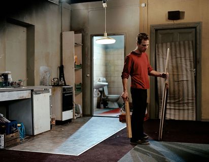In a run-down apartment stands a man in a red top with wooden strips in his hands.