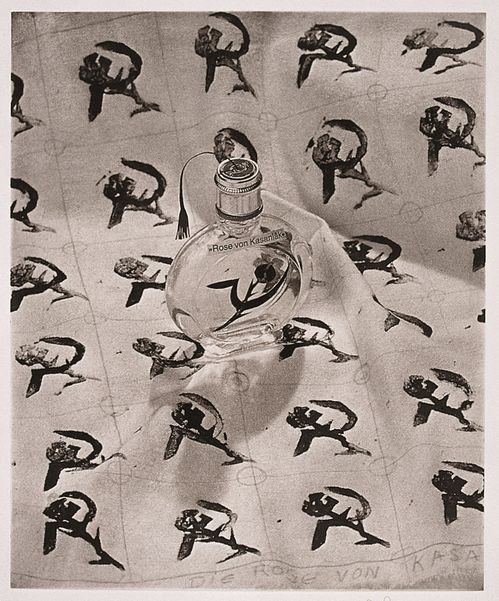 Die Photogravüre zeigt einen Flacon mit einer Lunte und der Beschriftung "Rose von Kasanlak", sowie eine Sichel mit einer Dornenrose. Der Flacon steht auf einem Papier, das mit einem Muster aus dem gleichen Motiv wiederkehrend bedeckt ist.