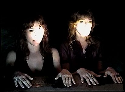 Two women are depicted halfway up the torso. Their hands are placed on a surface in front of them. Spotlights are directed at their faces.