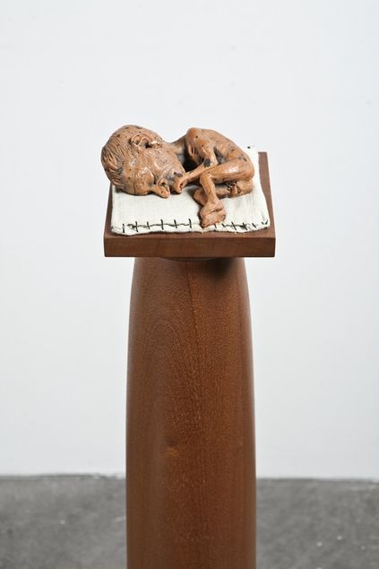 On a display mounted on a column lies a sculpture of a baby with an oversized head, its eyes closed, in a fetal position on a linen mat.