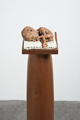 On a display mounted on a column lies a sculpture of a baby with an oversized head, its eyes closed, in a fetal position on a linen mat.