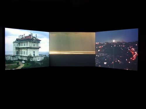 3 video projections side by side. On the left a three-storey villa, in the middle the horizon by the sea, an illuminated city in the dark and from a distance